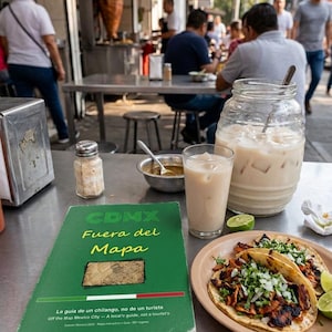 Puede incluir: Una guía verde titulada CDMX Fuera del Mapa sobre una mesa de metal junto a un plato con dos tacos de carne, cilantro y cebolla, un vaso de horchata, una jarra grande de horchata, un cuenco de salsa, un salero y rodajas de lima.