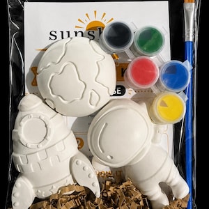 May include: A craft kit in a clear plastic bag containing three white ceramic figures: a planet, a rocket ship, and an astronaut. The kit includes five small circular paint pots in black, green, red, blue, and yellow, plus a blue-handled paintbrush.