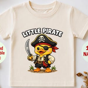 May include: A cream-coloured short-sleeve t-shirt featuring a graphic of a yellow duck dressed as a pirate with an eye patch, a black hat with a skull and crossbones, and a sword. The text LITTLE PIRATE is printed in black above the duck graphic.