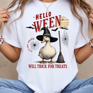 Könnte beinhalten: Ein weißes T-Shirt mit einer Grafik einer Ente, die einen schwarzen Hexenhut trägt, neben einem Besen. Der Text lautet HELLO WEEN, lets get spooky und WILL TRICK FOR TREATS. Das Design enthält einen gelben Mond, Spinnennetze und Fledermäuse.