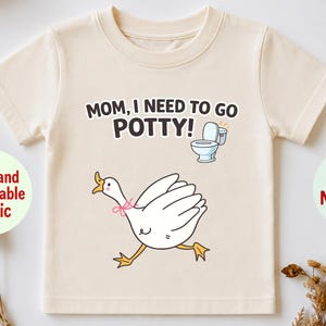 May include: A cream-colored short-sleeve t-shirt featuring a graphic of a white goose with a pink bow around its neck, a small toilet icon, and the black text "MOM, I NEED TO GO POTTY!" printed on the front center of the garment.