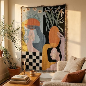 May include: A rectangular woven throw blanket featuring a split-panel design with two stylised figures holding large orange oval shapes above their heads, surrounded by white flowers, set against light blue and black backgrounds with a checkered base.