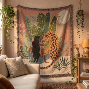 Pode incluir: Uma manta tecida com uma ilustração impressa de uma pessoa com cabelos longos e escuros sentada ao lado de um leopardo manchado entre grandes folhas tropicais verdes e uma lua pálida sobre um fundo rosa empoeirado com bordas franjadas.