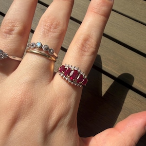 Könnte beinhalten: Ein Goldring mit fünf horizontal angeordneten, oval geschliffenen, tiefroten Rubinen, umgeben von einem Kranz aus kleinen, klaren Steinen. Daneben befinden sich ein schmaler Goldring und ein weiterer Goldring mit einem kleinen Mittelstein und einem strukturierten Design.