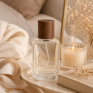 May include: A clear glass perfume bottle with a dark brown wooden cap. The front of the bottle features the name Olivia printed in gold cursive script. The bottle has a rectangular shape with rounded corners and a visible internal spray tube.