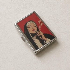 May include: A rectangular silver metal cigarette case featuring a graphic illustration of a nun wearing a black and white habit, smoking a cigarette with a thin trail of smoke rising against a solid red background. The case has a metal latch.
