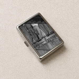 May include: A rectangular silver metal cigarette case featuring a black and white photograph on the lid. The image depicts a group of people standing in a line, viewed from the waist down, with a row of glass bottles positioned on a shelf below.