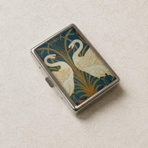 May include: A rectangular metal case featuring a decorative top panel with two white swans swimming among golden reeds against a deep teal background. The case has a polished silver-tone metal frame with a side latch mechanism for opening and closing.