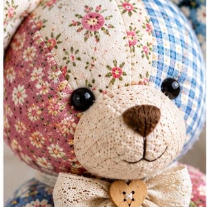 K&ouml;nnte beinhalten: Ein Stoffb&auml;r aus verschiedenen Stoffst&uuml;cken mit Blumenmustern, blauem Vichy-Karo und cremefarbenen Abschnitten. Der B&auml;r hat schwarze Kunststoffaugen, eine braun gestickte Nase und eine Spitzenschleife mit einem h&ouml;lzernen Herzknopf am Hals.