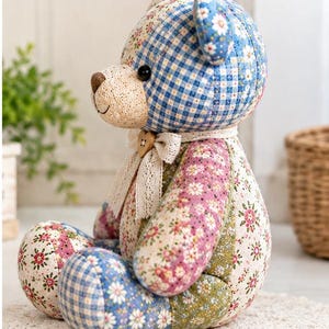 K&ouml;nnte beinhalten: Ein Stoffteddyb&auml;r aus verschiedenen Patchwork-Stoffen mit blauem Vichy-Muster, Blumenmustern in Rosa, Gr&uuml;n und Wei&szlig; sowie einer cremefarbenen Schnauze. Der B&auml;r tr&auml;gt ein Spitzenband um den Hals mit einem kleinen herzf&ouml;rmigen Holzknopf.