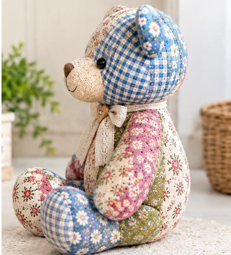 K&ouml;nnte beinhalten: Ein Stoffteddyb&auml;r aus verschiedenen Patchwork-Stoffen mit blauem Vichy-Muster, Blumenmustern in Rosa, Gr&uuml;n und Wei&szlig; sowie einer cremefarbenen Schnauze. Der B&auml;r tr&auml;gt ein Spitzenband um den Hals mit einem kleinen herzf&ouml;rmigen Holzknopf.