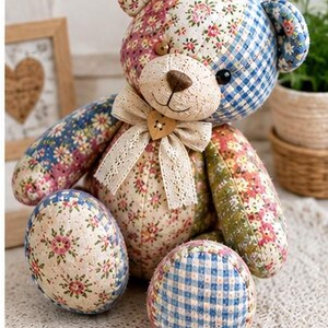 K&ouml;nnte beinhalten: Ein Stoffteddyb&auml;r aus verschiedenen gesteppten Stoffst&uuml;cken mit Blumenmustern und blauem Vichy-Karo. Der B&auml;r hat eine cremefarbene Schnauze, eine braun gestickte Nase, dunkle Augen und eine beige Spitzen-Schleife mit einem herzf&ouml;rmigen Knopf.