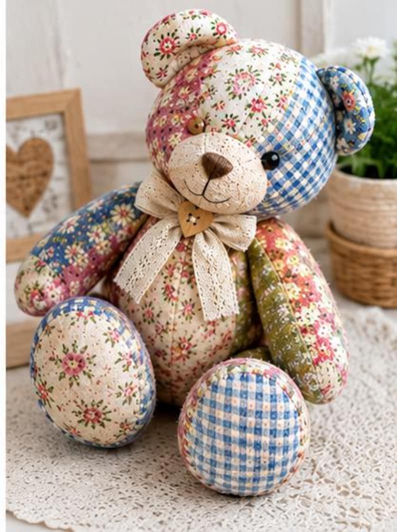K&ouml;nnte beinhalten: Ein Stoffteddyb&auml;r aus verschiedenen gesteppten Stoffst&uuml;cken mit Blumenmustern und blauem Vichy-Karo. Der B&auml;r hat eine cremefarbene Schnauze, eine braun gestickte Nase, dunkle Augen und eine beige Spitzen-Schleife mit einem herzf&ouml;rmigen Knopf.