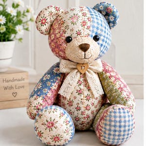 K&ouml;nnte beinhalten: Ein Patchwork-Teddyb&auml;r aus verschiedenen Stoffmustern, darunter rosa Blumen, blaues Vichy-Karo und blaue Blumenmuster. Der B&auml;r hat schwarze Knopfaugen, eine braun gestickte Nase und eine cremefarbene Spitzenschleife mit einem herzf&ouml;rmigen Holzknopf.