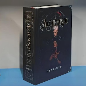 Op de afbeelding: Een hardcover boek getiteld Alchemised door Senlinyu met een zwarte kaft en een illustratie van twee figuren. De titel staat in gouden letters op de rug en voorkant, en de hoeken van het boek zijn verstevigd met zilverkleurige metalen accenten.