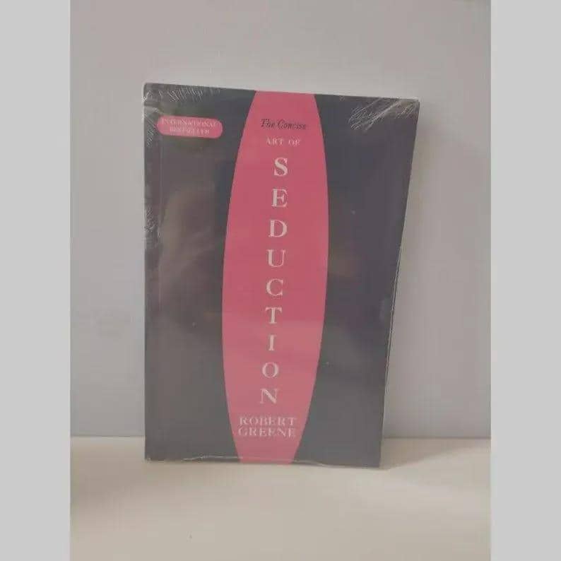 May include: A book titled The Concise Art of Seduction by Robert Greene, featuring a black cover with a large vertical pink oval in the center. The text is printed in white, and a small pink label reads International Bestseller on the top left.