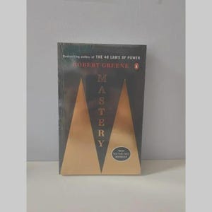 May include: A paperback book titled Mastery by Robert Greene, featuring a black cover with two large gold triangles and the text Bestselling author of THE 48 LAWS OF POWER. A circular sticker on the cover reads The #1 NEW YORK TIMES BESTSELLER.