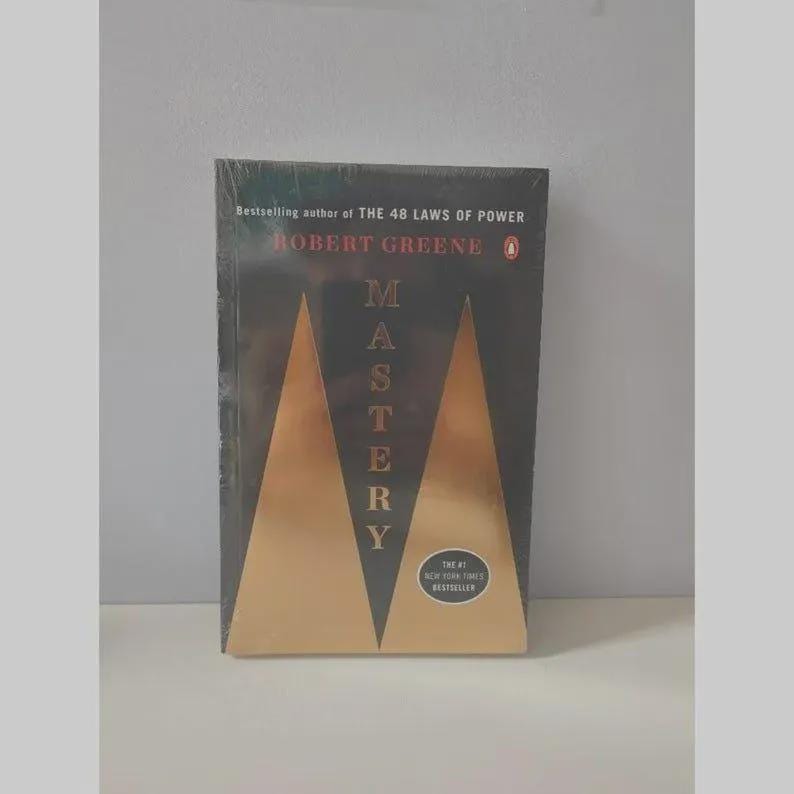 May include: A paperback book titled Mastery by Robert Greene, featuring a black cover with two large gold triangles and the text Bestselling author of THE 48 LAWS OF POWER. A circular sticker on the cover reads The #1 NEW YORK TIMES BESTSELLER.