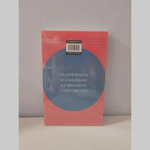 May include: A rectangular book with a coral-pink cover featuring a large dark blue circle in the center. The text inside the circle is upside down and reads: YOU ARE ABOUT TO BECOME AN APPRENTICE IN HUMAN NATURE. A barcode is at the top.