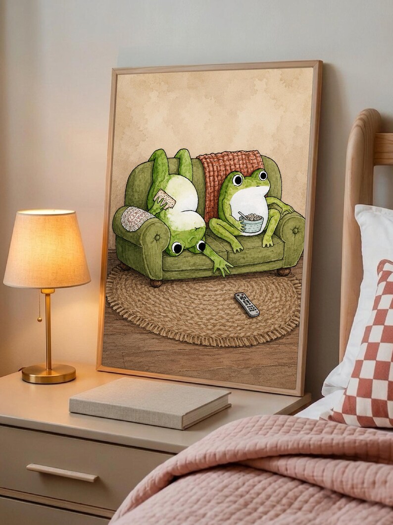 Puede incluir: Una l&aacute;mina art&iacute;stica enmarcada con la ilustraci&oacute;n de dos ranas verdes en un sof&aacute; verde. Una rana est&aacute; boca abajo sosteniendo un mando, mientras la otra est&aacute; sentada comiendo de un cuenco. Un mando a distancia descansa sobre una alfombra redonda.