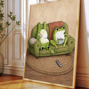 Puede incluir: Una l&aacute;mina de arte mural enmarcada con la ilustraci&oacute;n de dos ranas verdes en un sof&aacute; verde. Una rana est&aacute; boca abajo comiendo un aperitivo, mientras la otra est&aacute; sentada con un cuenco. Un mando a distancia descansa sobre una alfombra redonda tejida.