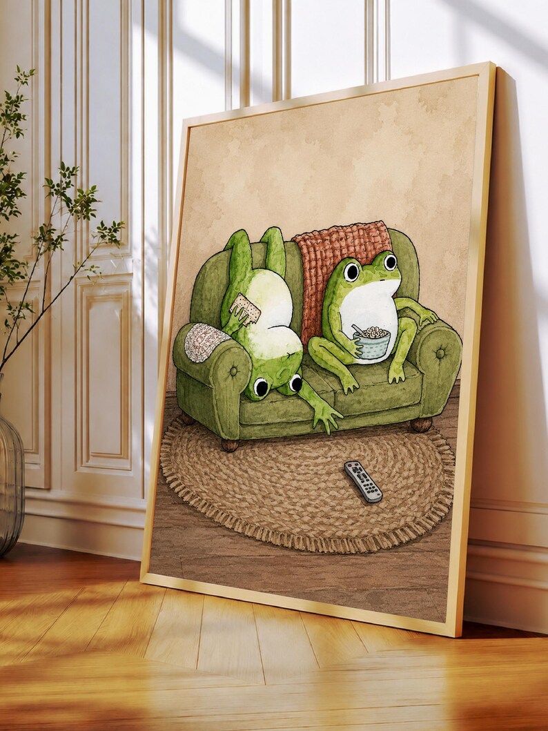 Puede incluir: Una l&aacute;mina de arte mural enmarcada con la ilustraci&oacute;n de dos ranas verdes en un sof&aacute; verde. Una rana est&aacute; boca abajo comiendo un aperitivo, mientras la otra est&aacute; sentada con un cuenco. Un mando a distancia descansa sobre una alfombra redonda tejida.