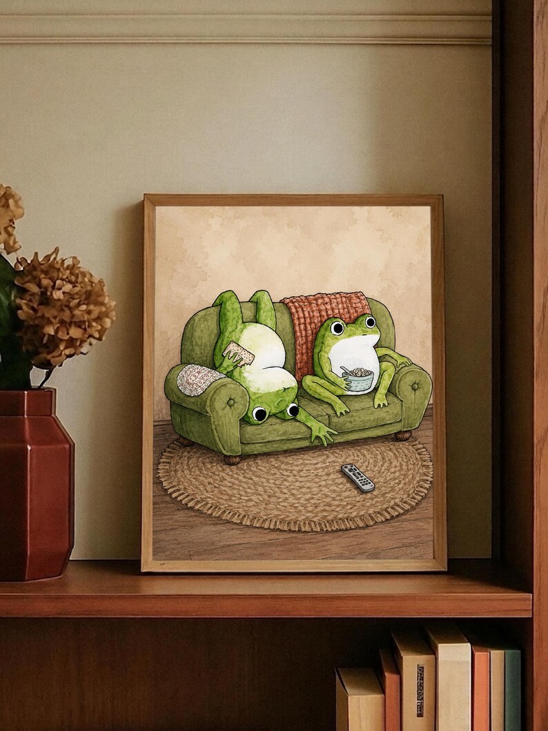 Puede incluir: Una l&aacute;mina de arte mural enmarcada que muestra dos ranas verdes en un sof&aacute; verde. Una rana est&aacute; boca abajo comiendo un aperitivo, mientras la otra est&aacute; sentada con un cuenco. Un mando a distancia est&aacute; sobre una alfombra redonda frente al sof&aacute; y una manta roja cuelga del respaldo.
