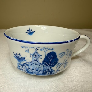 May include: A white ceramic teacup featuring a blue transferware pattern of a pagoda, trees, and a boat on the exterior. The rim is accented with a thin blue line, and a small blue boat illustration is visible on the interior wall of the cup.