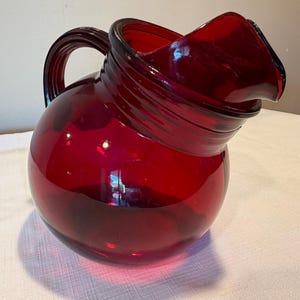 May include: A deep red glass pitcher with a spherical body, a wide pouring spout, and a curved handle. The glass features a translucent finish with horizontal ribbed detailing around the neck area, creating a smooth, rounded silhouette.