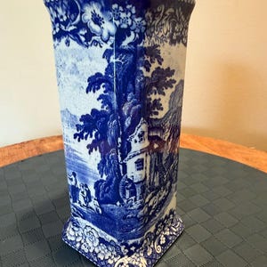 May include: A square-shaped ceramic vase featuring a blue and white transferware pattern. The design depicts a detailed landscape with trees, a house, and figures. The top and bottom edges are decorated with a floral border, and the surface has a visible crackle glaze finish throughout the entire piece.