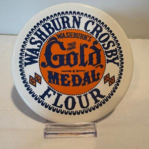 Könnte beinhalten: Ein runder weißer Keramikt weißer Keramikt weißer Keramikteller mit einer Vintage-Werbung für Washburn Crosby Gold Medal Mehl. Das Design umfasst blauen Text, einen orangefarbenen Mittelkreis und zwei kleine orangefarbene Rautenlogos mit den Buchstaben CC.