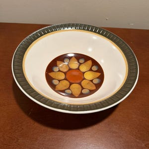 Puede incluir: Un cuenco de cerámica redondo con interior color crema y borde acanalado de color verde oscuro. El centro del cuenco presenta un diseño circular con un núcleo naranja sólido rodeado por un anillo de patrones amarillos en forma de pétalo sobre un fondo marrón oscuro.