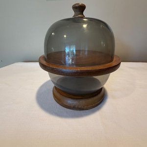 May include: A round wooden serving stand featuring a dark brown wood base and a matching wooden rim. The stand includes a removable, translucent dark gray glass dome cover with a small wooden knob handle at the top for easy lifting and access.