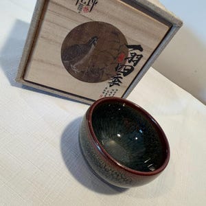 Może przedstawiać: Mała, okrągła ceramiczna miseczka z ciemnym, nakrapianym szkliwem, głębokim czerwonym brzegiem i teksturowanym, spękanym wzorem wewnątrz. Za miseczką znajduje się kwadratowe, jasne drewniane pudełko z wieczkiem ozdobionym ilustracją ptaka.