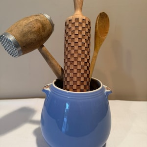 May include: A light blue ceramic crock with two small handles containing a wooden meat mallet with a metal head, a wooden rolling pin with a checkered pattern, and a wooden spoon. The kitchen tools are arranged upright inside the blue container.