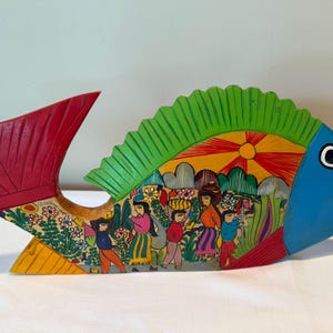 May include: A hand-painted wooden fish sculpture featuring a vibrant red tail, a bright green dorsal fin, and a blue head. The body displays a detailed scene of people in a landscape with a yellow sun, colorful flowers, and green foliage.