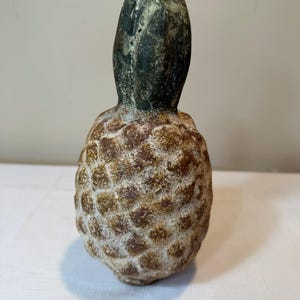 May include: A decorative pineapple-shaped object with a weathered, textured surface. The body features a brown, mottled finish with a raised, diamond-patterned texture, while the top section is a dark, muted green with a smooth, rounded shape.