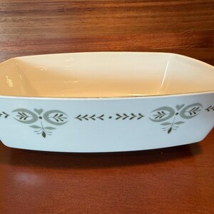 May include: A rectangular white ceramic serving dish with rounded corners and a tapered base. The exterior features a repeating sage green botanical pattern consisting of stylized leaves, small hearts, and dots arranged in a horizontal band.
