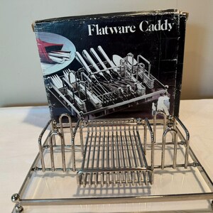 May include: A silver-toned metal flatware caddy featuring a rectangular base with a central grid compartment and four upright dividers for organizing utensils. The product is displayed in front of its original black cardboard packaging box.