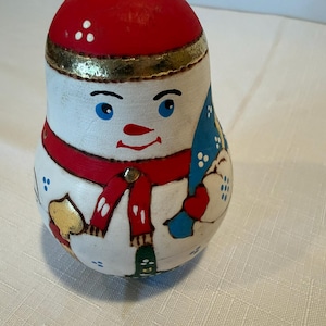 May include: A hand-painted wooden nesting doll shaped like a snowman with a red hat featuring white dots, a gold band, blue eyes, and a red scarf. The snowman holds a blue object and has a gold-colored accent on the side, with a white base.