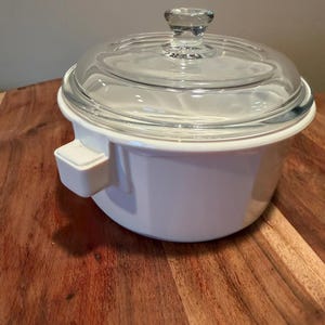 Puede incluir: Una fuente de cerámica blanca para horno con tapa de cristal transparente. El recipiente tiene un cuerpo redondo con base plana y un pequeño asa cuadrada que sobresale por el lateral. La tapa de cristal tiene un pomo central.