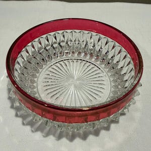 May include: A round glass bowl featuring a clear, diamond-cut pattern on the sides and a sunburst design on the base. The upper rim is accented with a smooth, solid band of deep red glass, contrasting with the transparent, faceted body of the bowl.