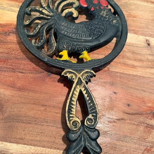 Può includere: Un sottopentola circolare in ghisa raffigurante un gallo nero con cresta rossa e zampe gialle. Il design include un manico ornato con accenti color oro e volute intricate. Il gallo è raffigurato di profilo con piume dettagliate.