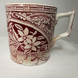 May include: A ceramic mug featuring a deep red and cream transferware pattern. The design displays intricate floral motifs, scrolling foliage, and geometric borders. The mug has a sturdy, angular handle and a visible crackle glaze finish.
