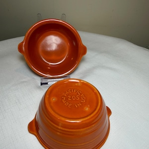 May include: Two orange ceramic bowls with side handles. One bowl is positioned upright, while the other is inverted to show the base, which is embossed with the text PACIFIC 205 MADE IN USA. The bowls feature a smooth, glossy finish and circular ridges.