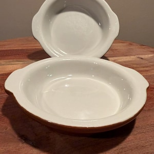 May include: Two round ceramic baking dishes with white interiors and brown exteriors. Each dish features two small, integrated side handles and a smooth, glossy finish. The dishes are stacked slightly, with one positioned behind the other.