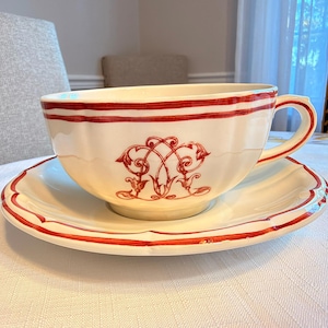 May include: A cream-colored ceramic teacup and saucer set featuring a scalloped edge and hand-painted red trim. The cup displays a decorative red monogram design on the front, with matching red lines circling the rim and saucer edge.