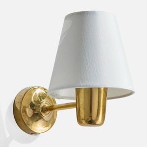 Può includere: Una lampada da parete con base circolare in ottone lucido, un sottile braccio orizzontale in ottone e un portalampada cilindrico in ottone. L'apparecchio è completato da un paralume in tessuto bianco di forma conica.