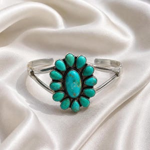 May include: A silver cuff bracelet featuring a large oval turquoise stone at the centre, surrounded by twelve smaller teardrop-shaped turquoise stones arranged in a floral pattern. The metal band is smooth and polished with a split design.