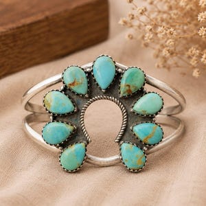 May include: A silver cuff bracelet featuring a central horseshoe-shaped frame adorned with nine teardrop-shaped turquoise stones. The stones display varying shades of blue and green with brown matrix patterns, set against a dark oxidised silver base.